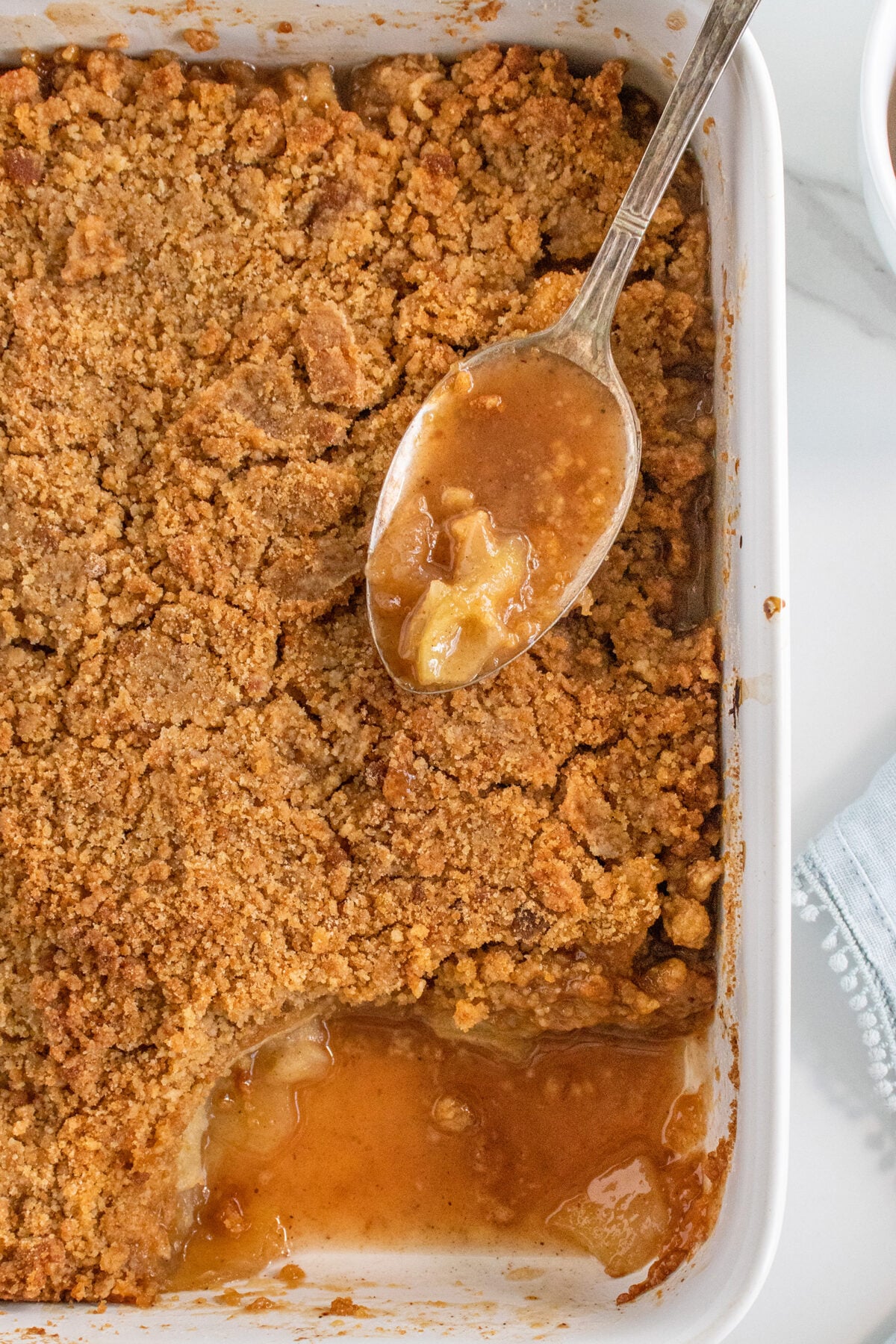 A white baking dish filled with apple crisp; part of the dessert has been scooped out, revealing baked apple filling and a golden brown crumbly topping. A spoon rests in the dish with apple filling on it.