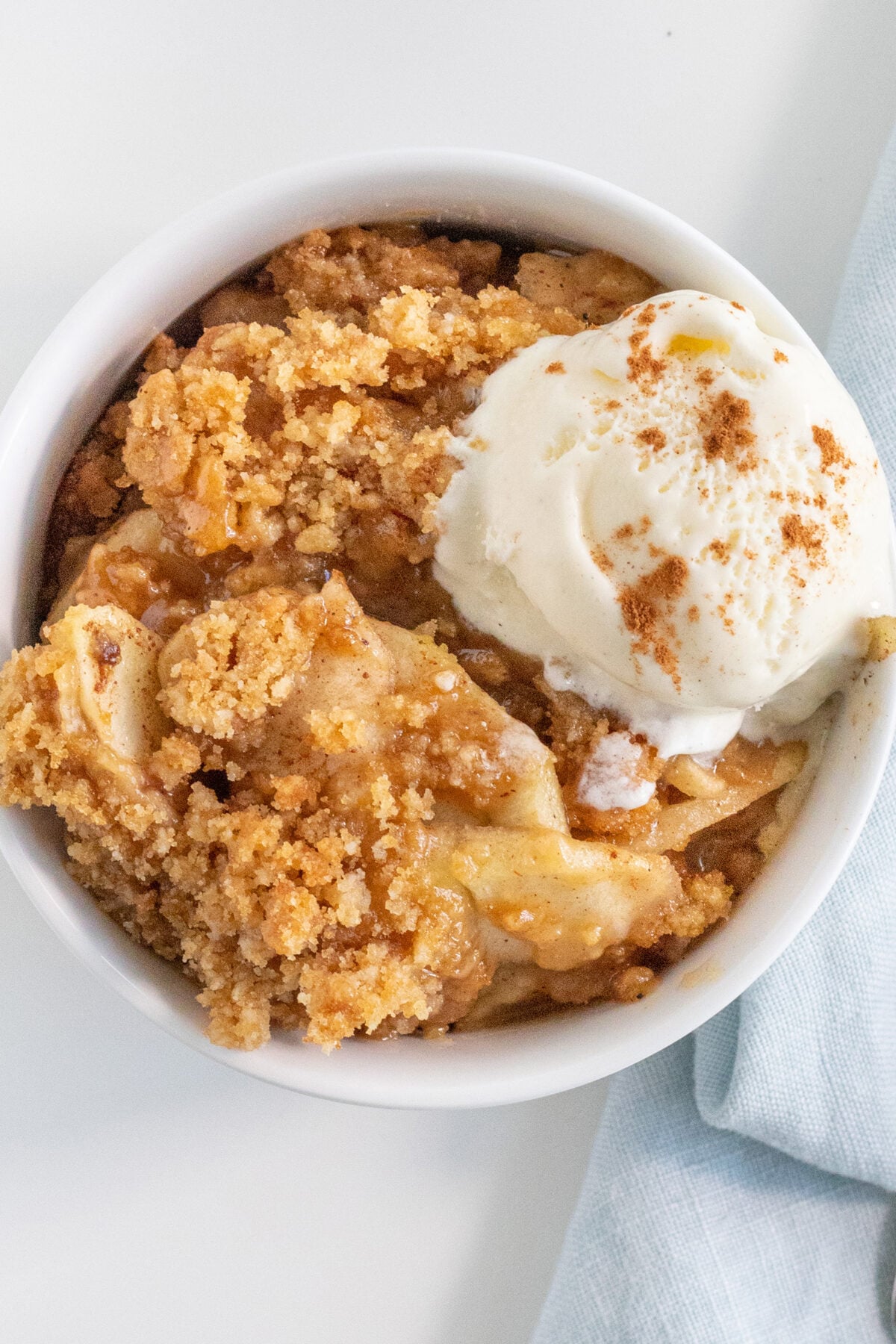 A bowl of apple crumble topped with a scoop of vanilla ice cream and a sprinkle of cinnamon, set on a white surface next to a light blue cloth.