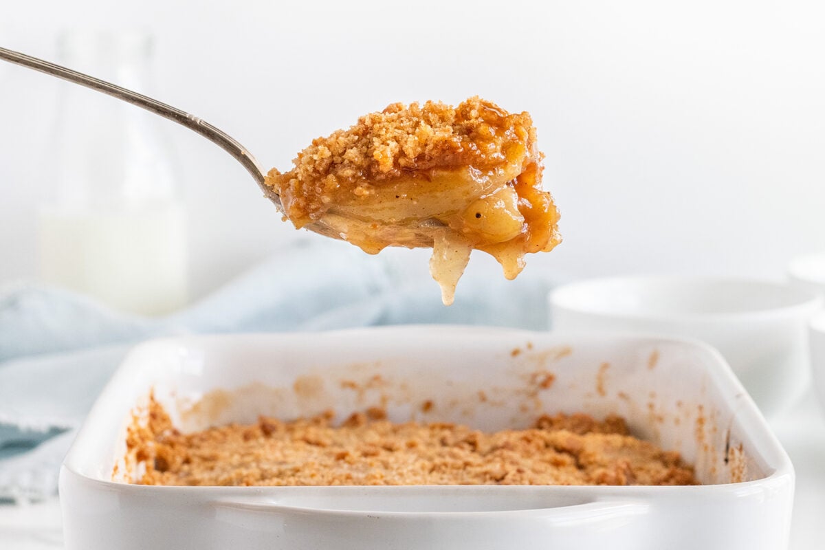 A spoonful of golden-brown apple crumble held above a white baking dish filled with the dessert, with part of the crumble missing. The background is softly blurred with cups and a milk bottle.