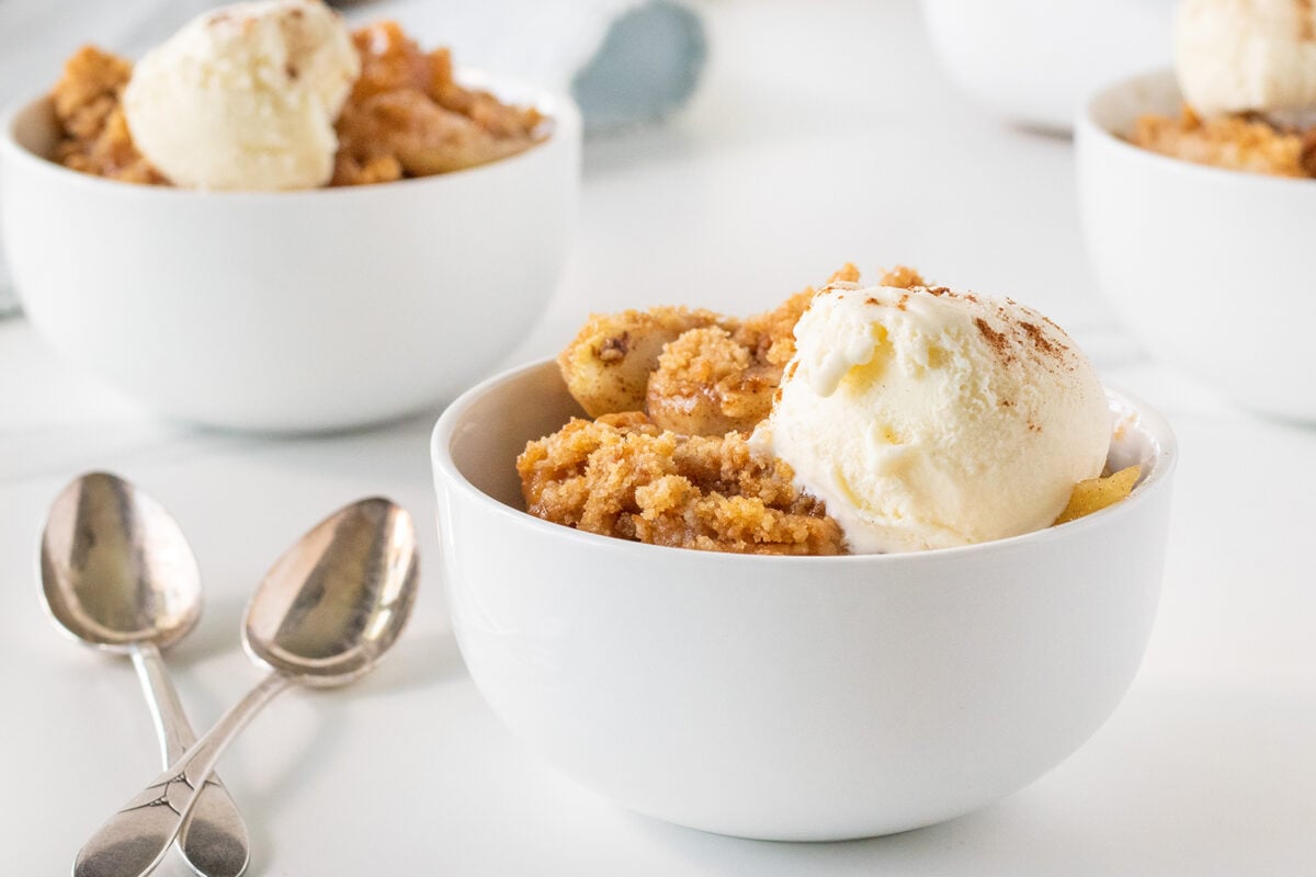 A white bowl filled with apple crumble and a scoop of vanilla ice cream, next to two metal spoons. In the background, there are more bowls with the same dessert on a white surface.