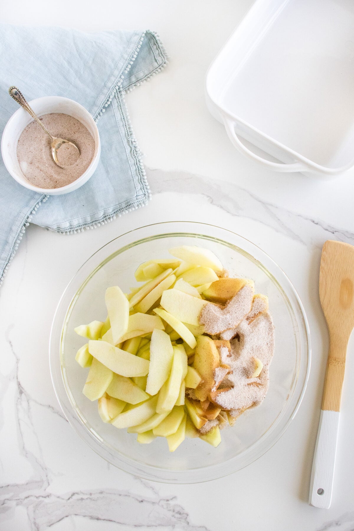 A glass bowl with peeled apple slices sprinkled with cinnamon sugar sits on a white marble surface. Nearby are a wooden spatula, a white baking dish, and a small bowl of cinnamon sugar with a spoon on a light blue napkin.