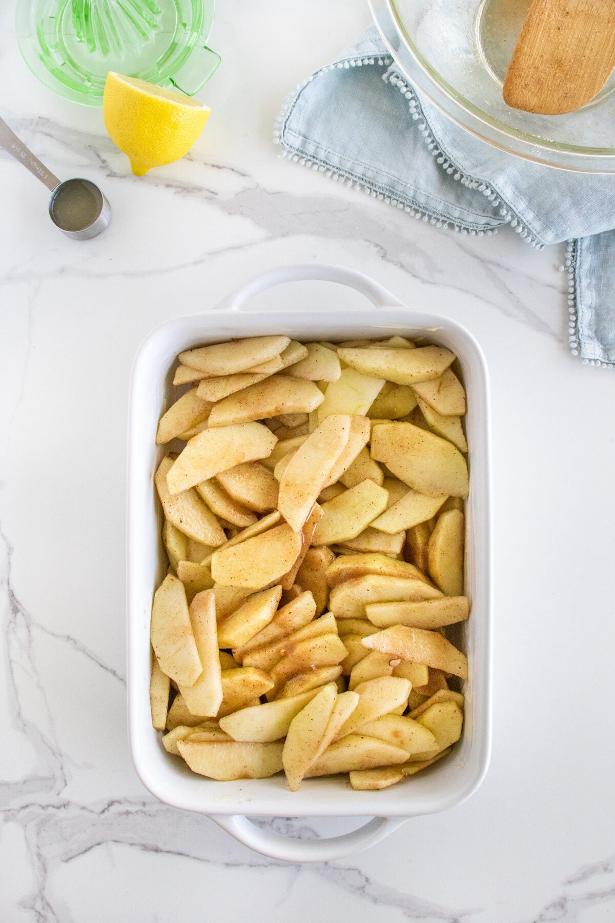 A white baking dish filled with sliced apples coated in cinnamon sits on a marble countertop. Nearby are a lemon wedge, a green citrus juicer, a metal measuring spoon, and a blue cloth napkin.