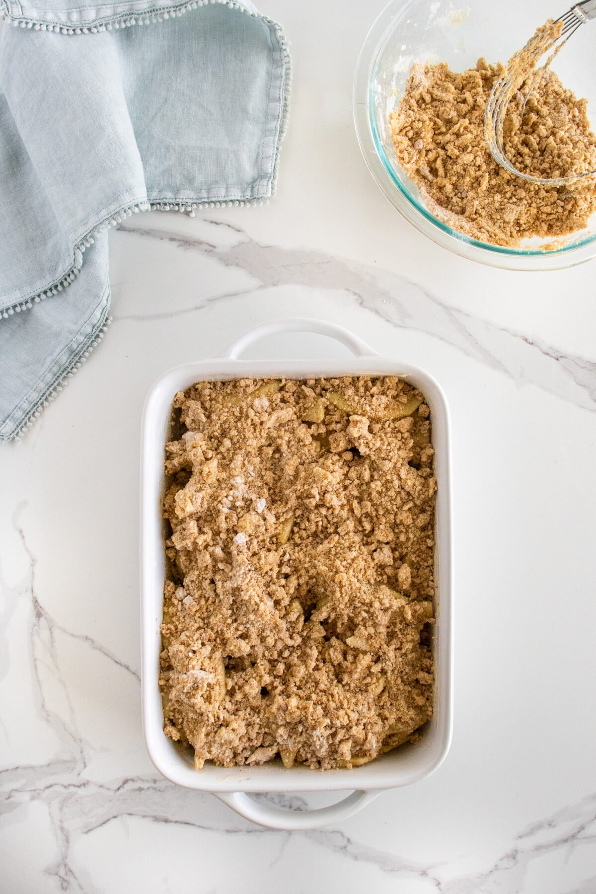 A white baking dish filled with an unbaked crumbly mixture sits on a marble surface next to a glass bowl with remaining crumb topping and a light blue cloth napkin.