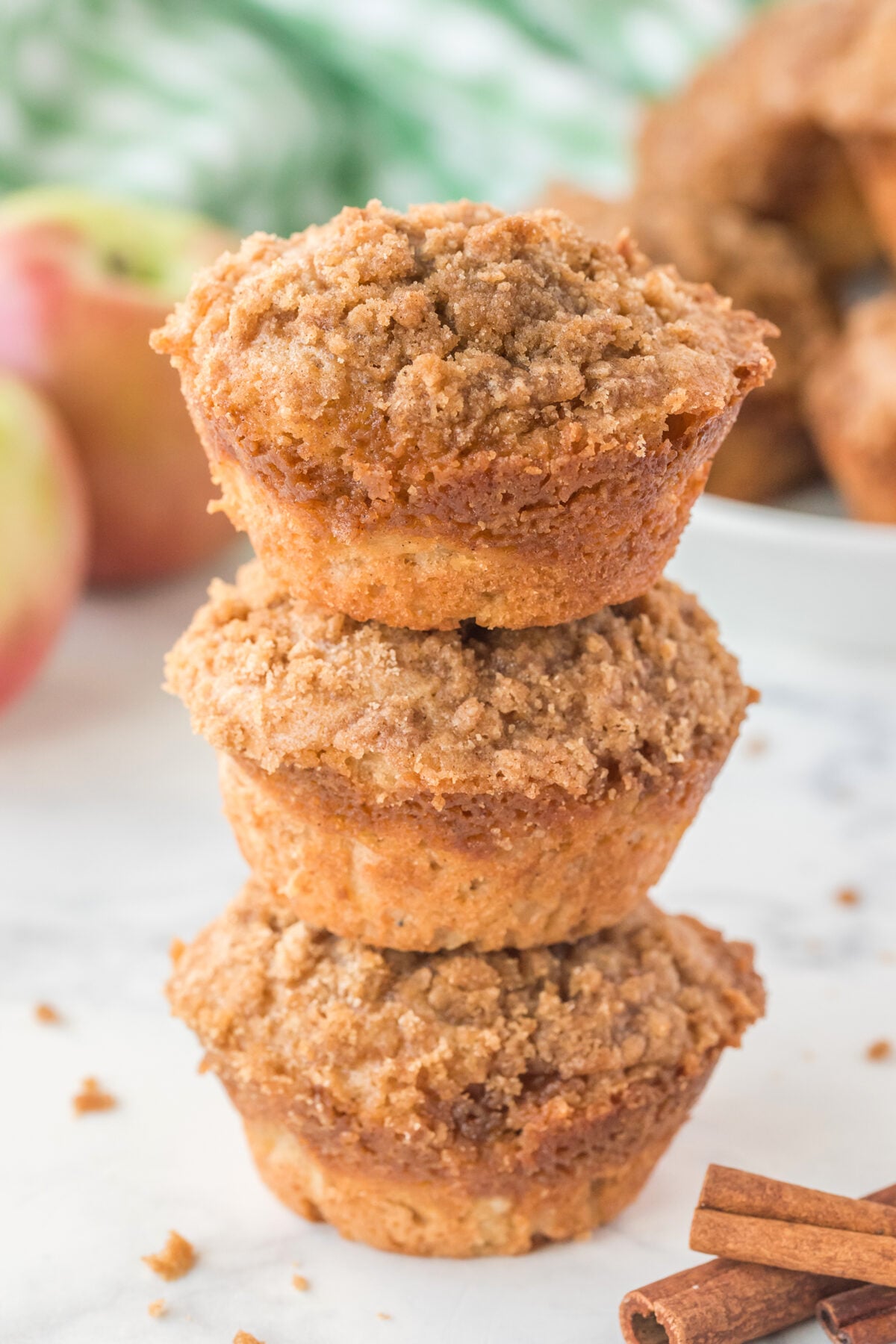 Three crumb-topped muffins are stacked on top of each other on a light surface, with apples and cinnamon sticks in the background. The muffins have a golden, crumbly topping.