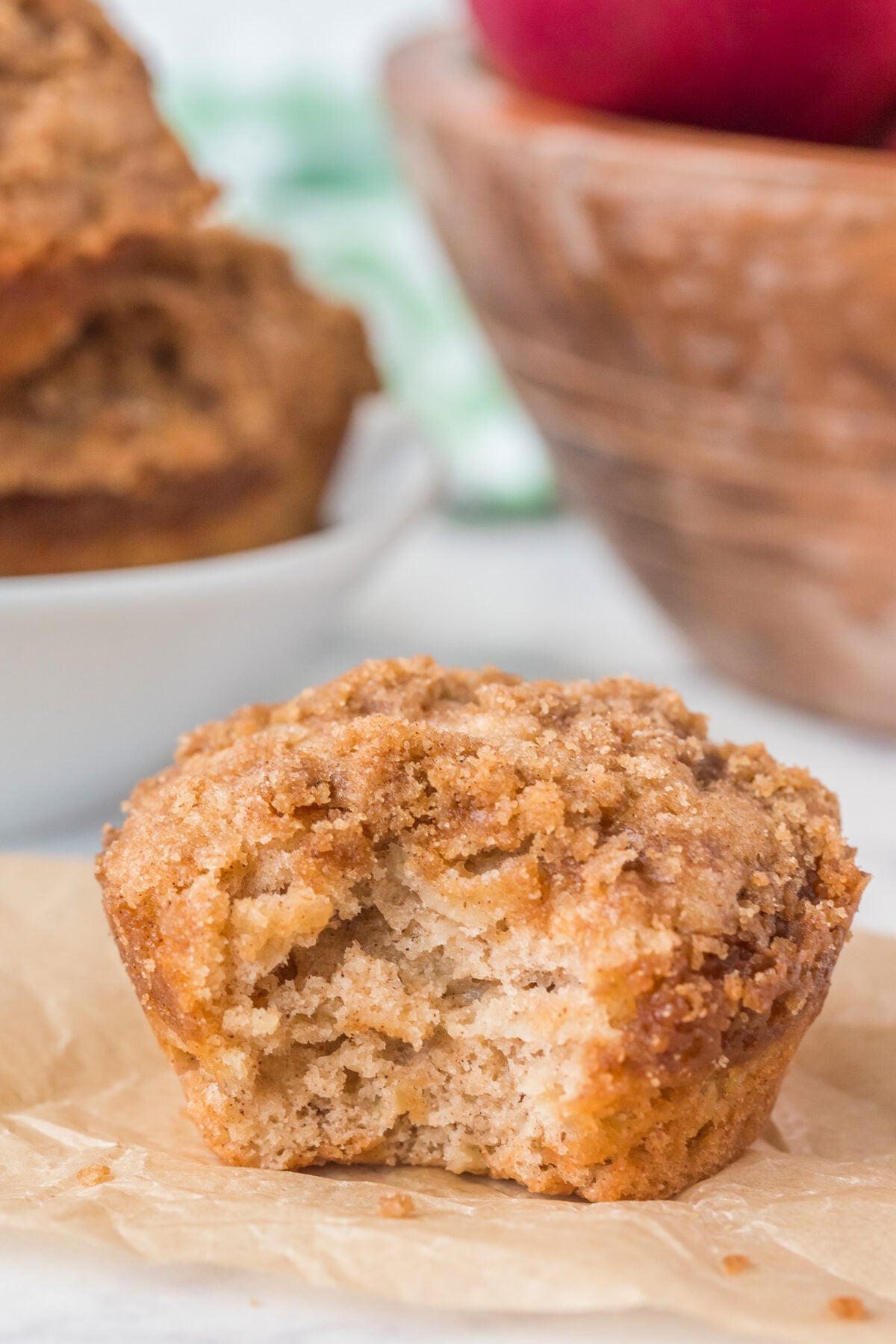 A close-up of a crumb-topped muffin with a bite taken out, resting on parchment paper. In the background, there’s a bowl of more muffins and a wooden bowl with red apples.