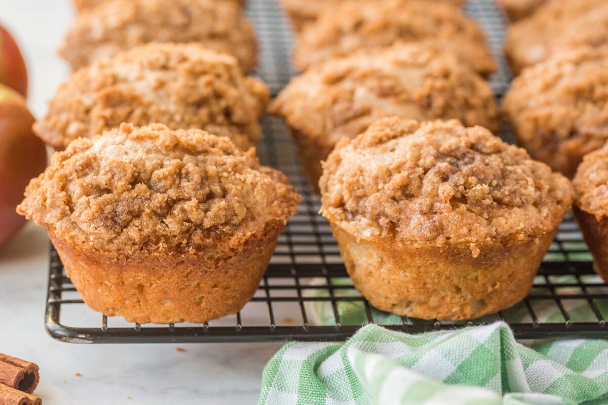 Close-up of freshly baked muffins with a crumbly topping cooling on a wire rack, with a green and white checkered cloth and cinnamon sticks nearby.