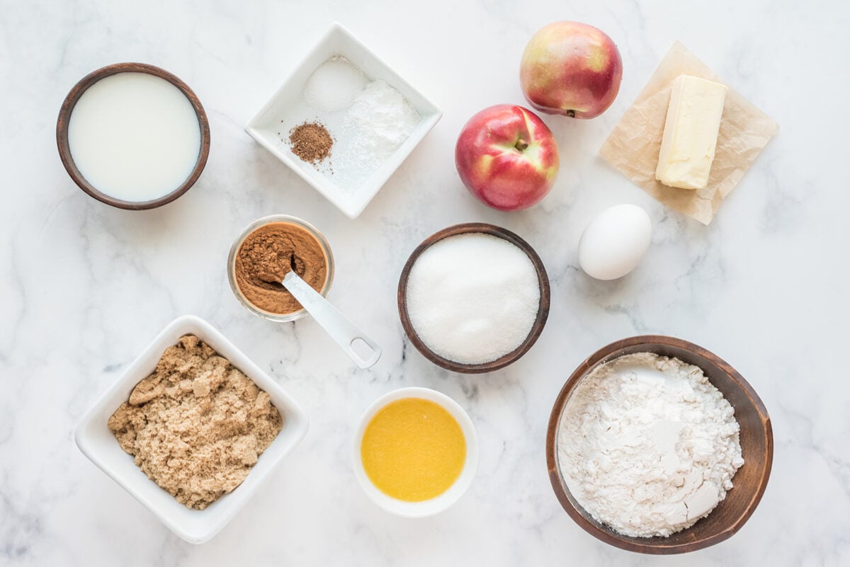 A top-down view of baking ingredients on a white marble surface, including milk, sugar, flour, brown sugar, spices, baking powder, salt, an egg, butter, melted butter, and two peaches.