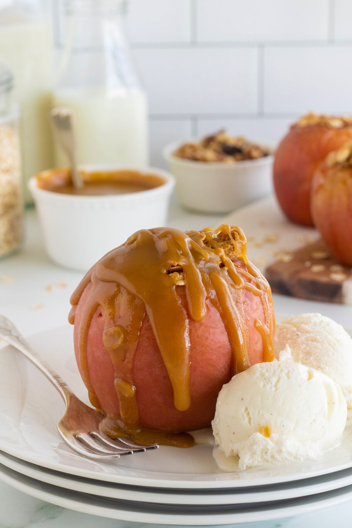 A baked apple drizzled with caramel sauce sits on a plate with a scoop of vanilla ice cream. A fork rests beside the dessert, and ingredients like caramel sauce, granola, and whole apples are in the background.