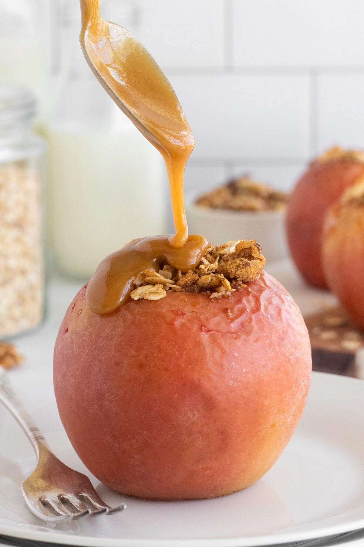 A spoon drizzles caramel sauce over a baked apple topped with oat crumble, placed on a white plate with a fork beside it. More baked apples and ingredients are blurred in the background.