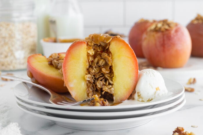 A halved baked peach stuffed with oat crumble sits on a white plate next to a scoop of vanilla ice cream. A fork rests on the plate, and more whole peaches and a jar of oats are in the background.