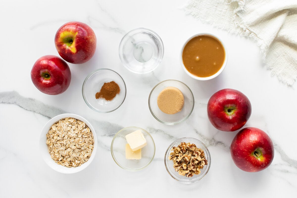 Overhead view of five red apples, oats, butter, brown sugar, walnuts, cinnamon, a bowl of caramel sauce, and a small bowl of water arranged neatly on a white marble surface near a cream-colored cloth.