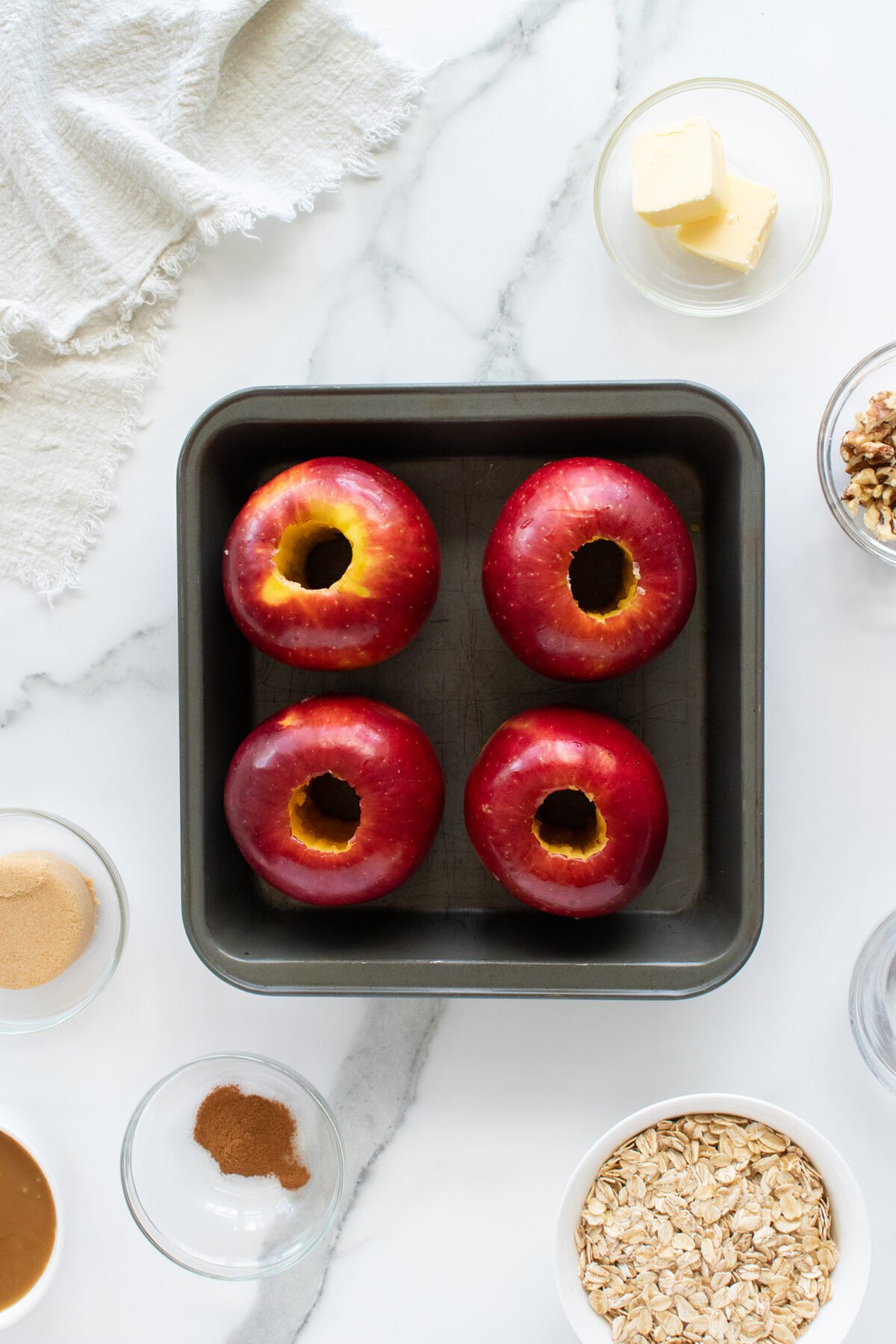 A baking pan with four cored red apples sits on a marble surface, surrounded by small bowls of oats, brown sugar, walnuts, cinnamon, butter, and a white cloth in the corner.