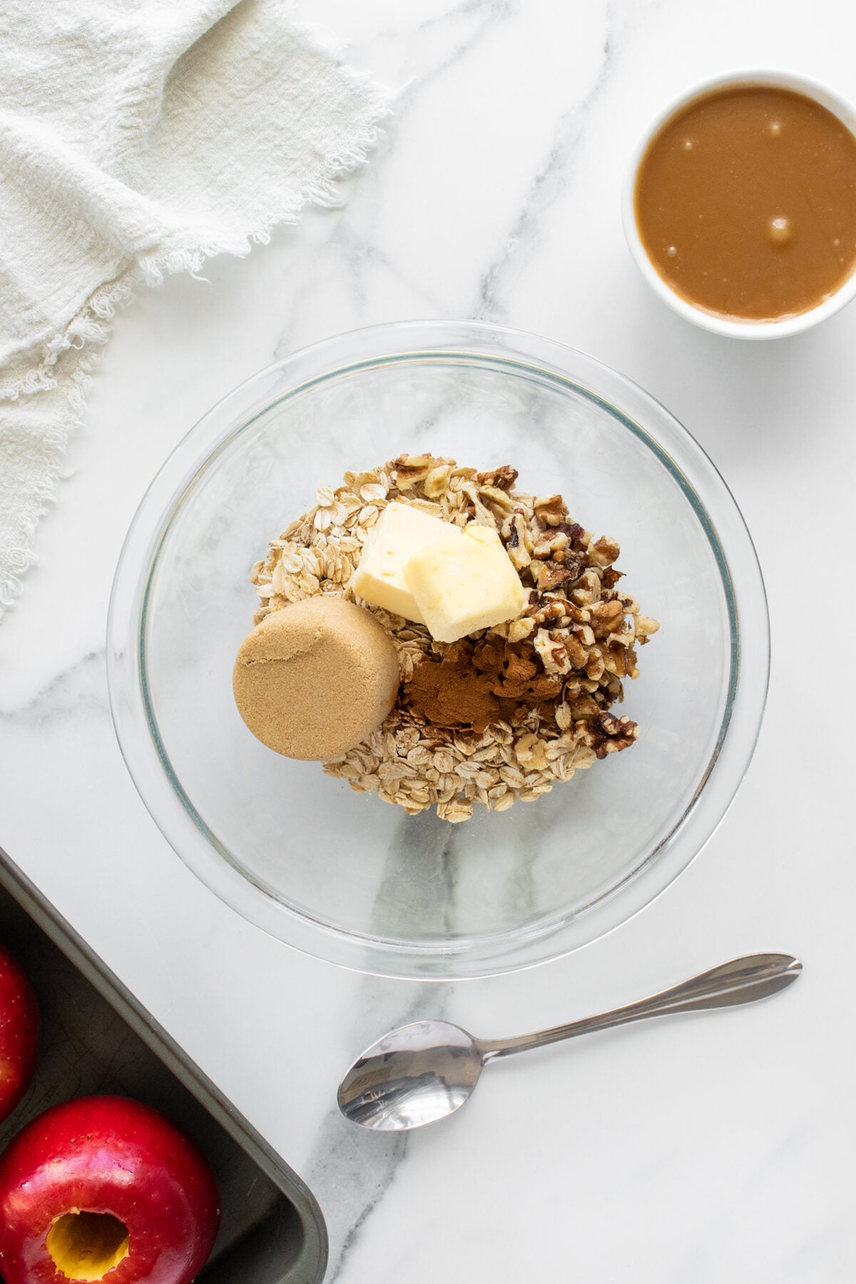 A glass bowl with oats, brown sugar, butter, cinnamon, and nuts sits on a marble surface. Nearby are a spoon, a cup of caramel, a baking tray with red apples, and a white cloth.