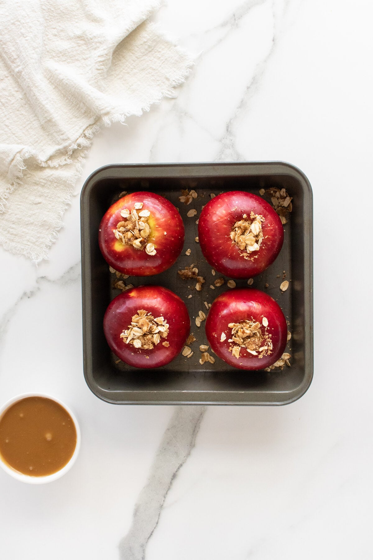 A square metal baking dish with four red apples filled with an oat mixture sits on a white marble surface. A small white bowl of caramel sauce and a beige cloth are nearby.