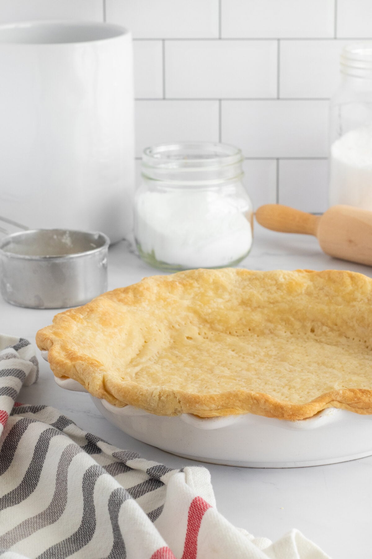 A baked pie crust in a white pie dish sits on a counter with a striped towel, a metal measuring cup, a jar of flour, and a rolling pin in the background. White subway tiles are on the wall.