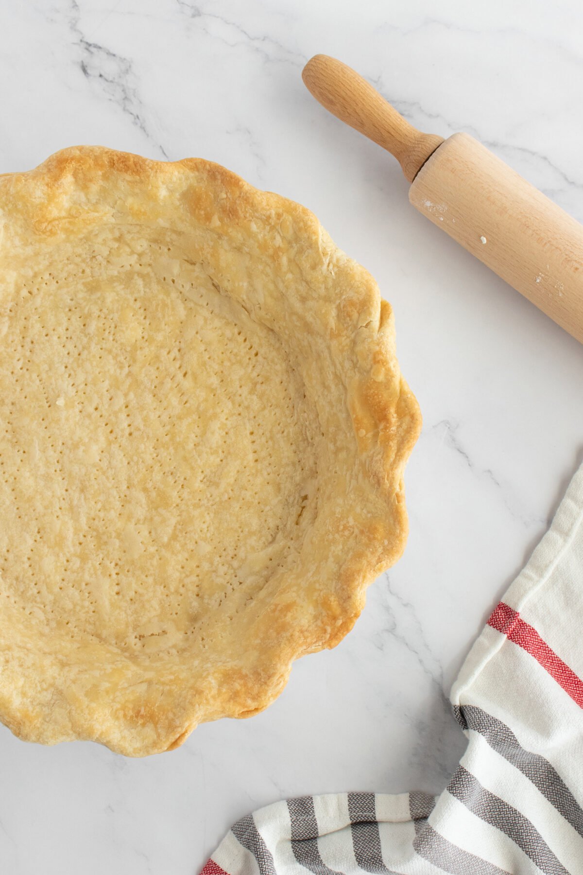 A baked pie crust in a fluted pie dish sits on a marble surface next to a wooden rolling pin and a striped kitchen towel.