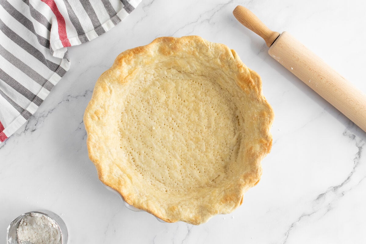 A baked pie crust in a fluted pie dish on a marble surface, with a rolling pin, a striped kitchen towel, and a small bowl of flour nearby.