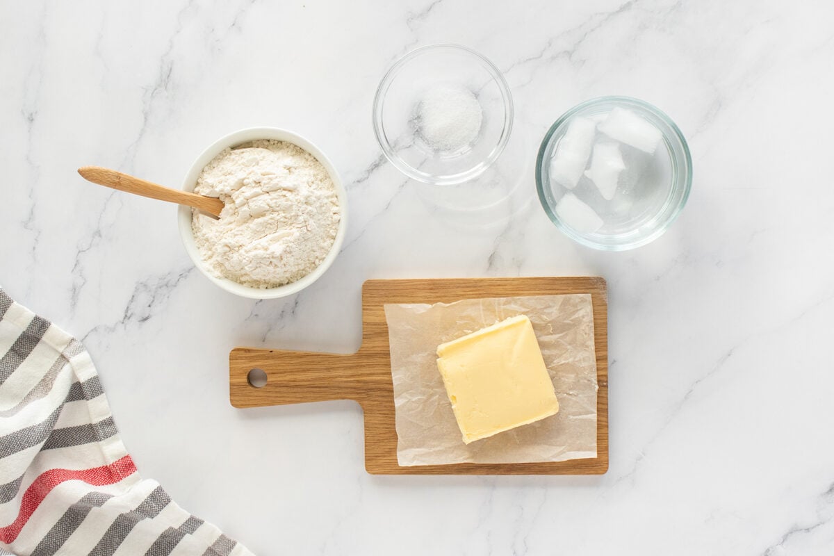 A bowl of flour with a wooden spoon, a glass of water, a glass of salt, and a block of butter on a small wooden board, all arranged on a white marble surface next to a striped cloth.