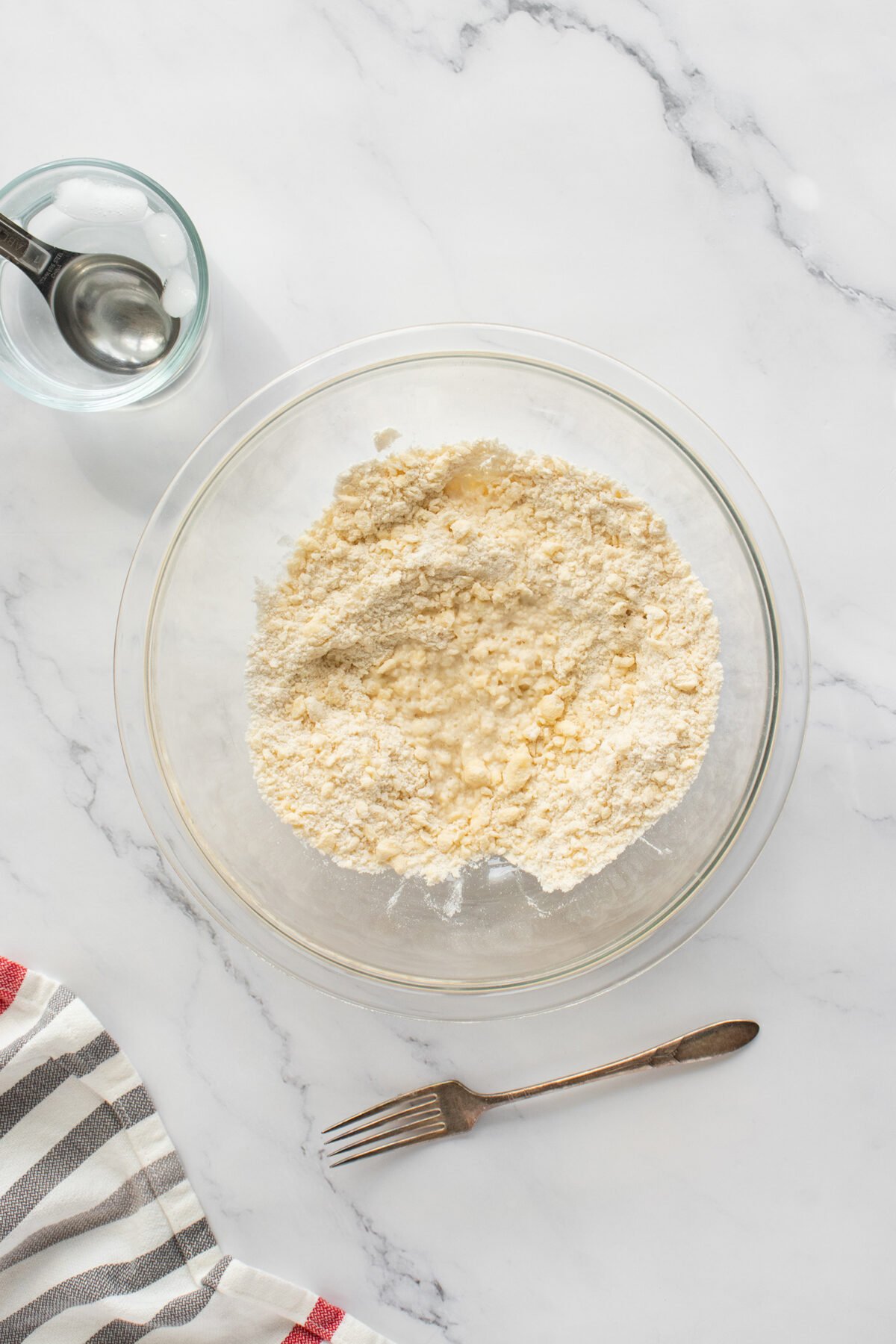 A glass bowl filled with dry baking ingredients on a marble countertop, beside a metal fork, a striped kitchen towel, and a small measuring cup with water.