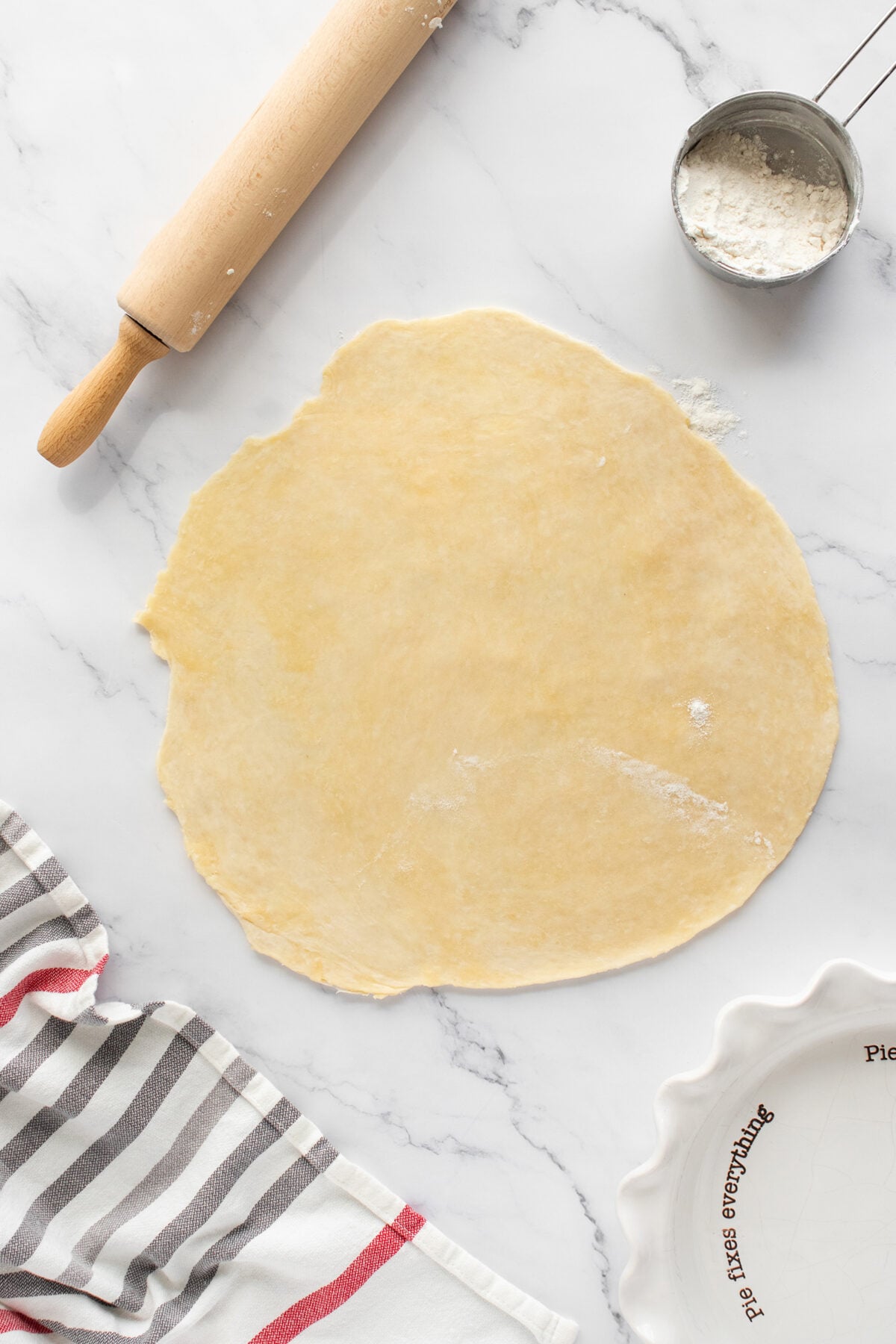 A round sheet of rolled-out dough on a marble surface, with a wooden rolling pin, a metal cup of flour, a striped towel, and a white pie dish nearby.