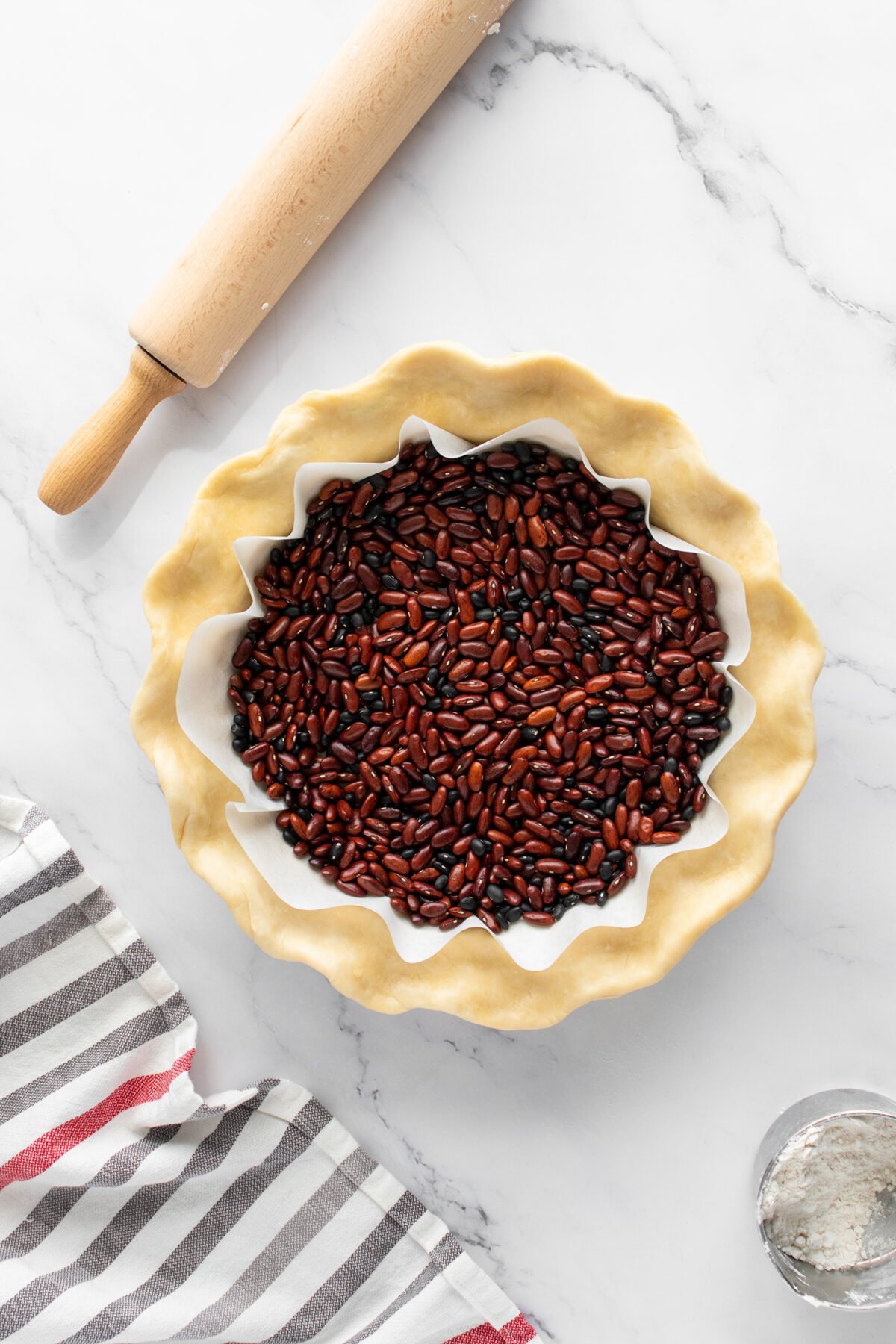A pie crust in a fluted dish is lined with parchment paper and filled with dried beans for blind baking. A rolling pin, a striped kitchen towel, and a small bowl of flour are nearby on a marble surface.