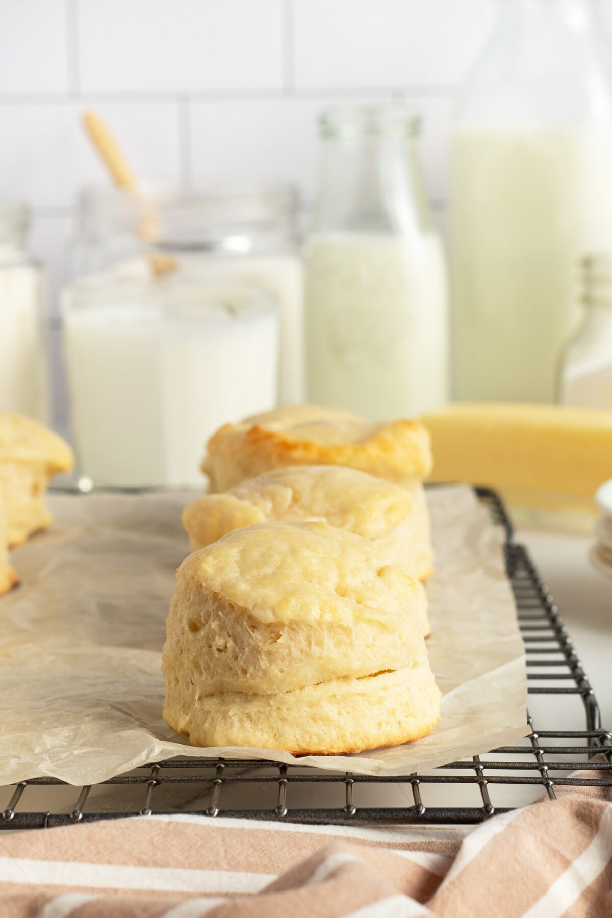 Freshly baked biscuits rest on a parchment-lined cooling rack, with jars and bottles of milk in the background on a kitchen counter. A beige-striped kitchen towel is partially visible at the bottom.