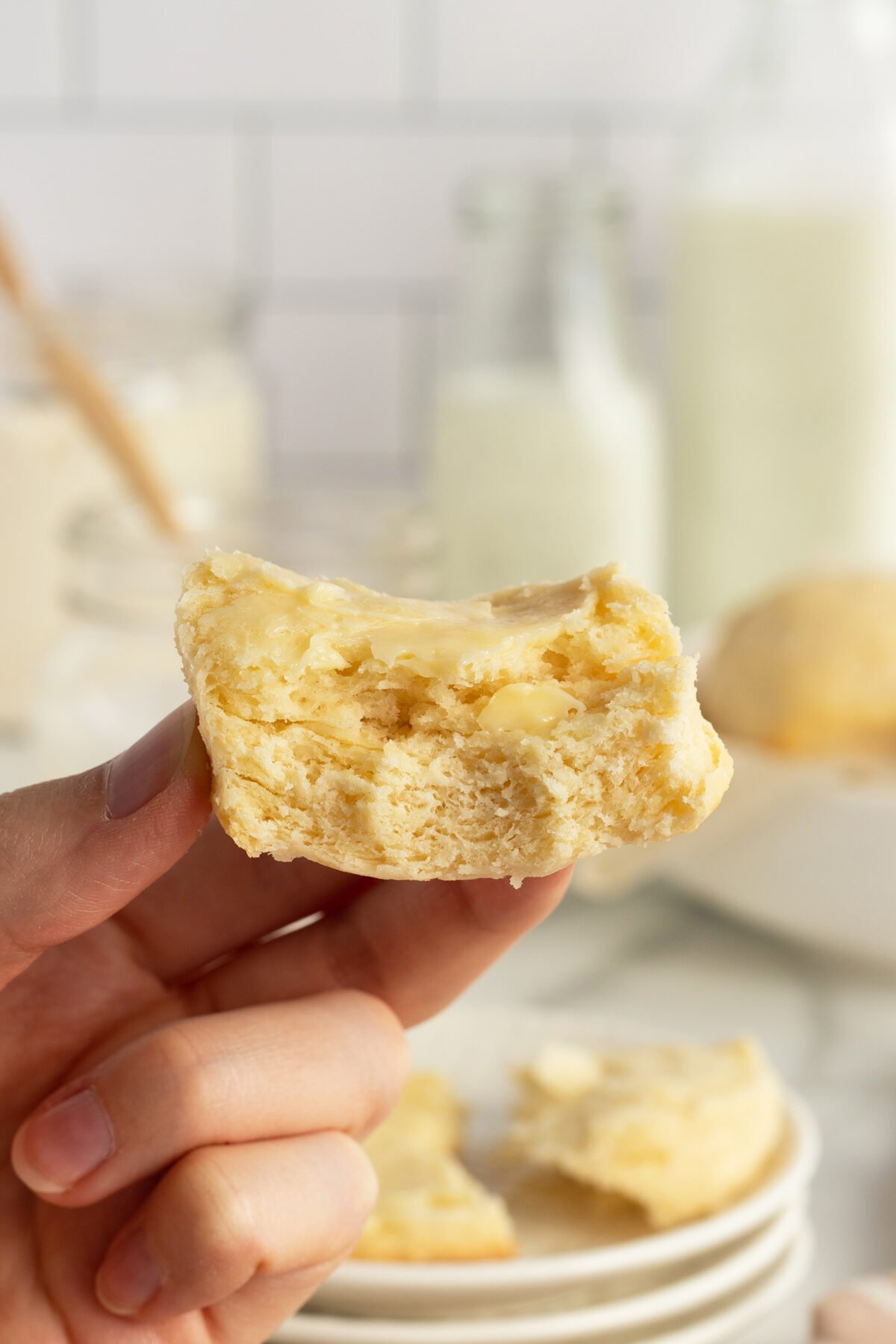 A hand holds a half-eaten biscuit with melting butter on top, with milk bottles and another biscuit blurred in the background.