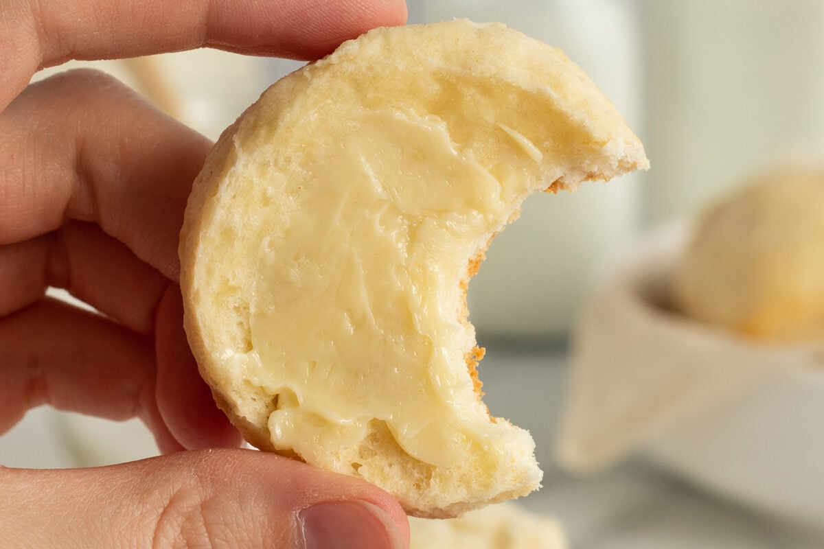 A hand holds a biscuit with a bite taken out, showing a layer of melted butter spread on top.