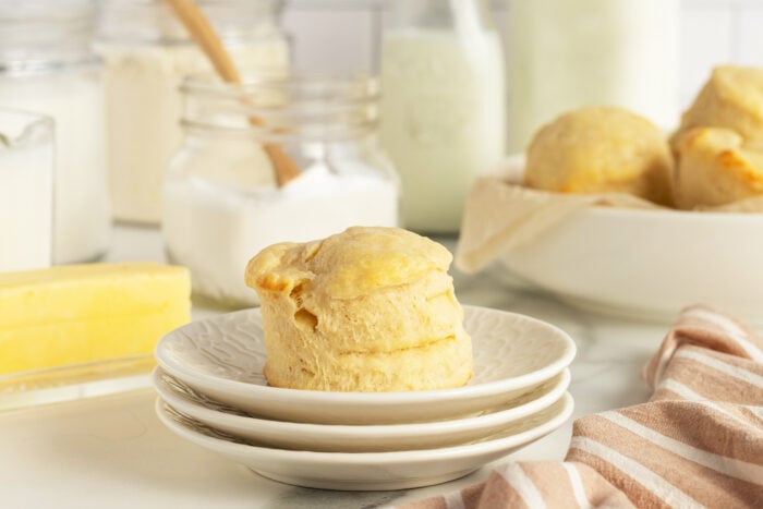 A golden biscuit sits on stacked white plates, surrounded by baking ingredients like butter, flour, and milk in jars, with more biscuits in a bowl in the background and a striped cloth in the foreground.