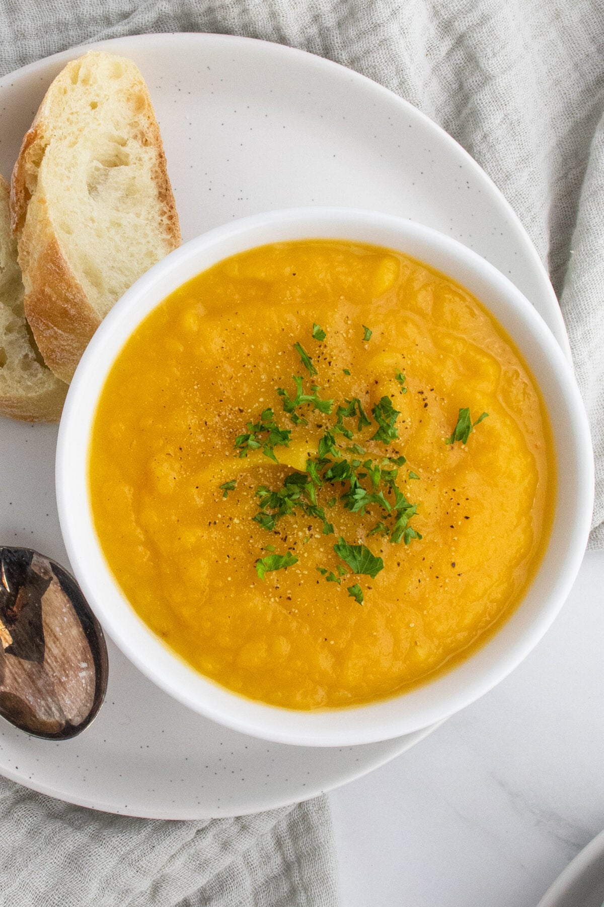 A white bowl of orange-colored soup garnished with chopped herbs and black pepper, served with a slice of bread on a white plate, next to a wooden spoon.
