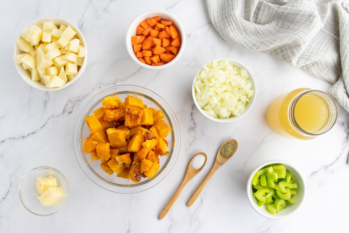A flat lay of bowls containing chopped potatoes, carrots, onions, celery, cooked squash, butter, and a jar of broth on a white marble surface, with salt and pepper in wooden spoons and a gray cloth in the corner.