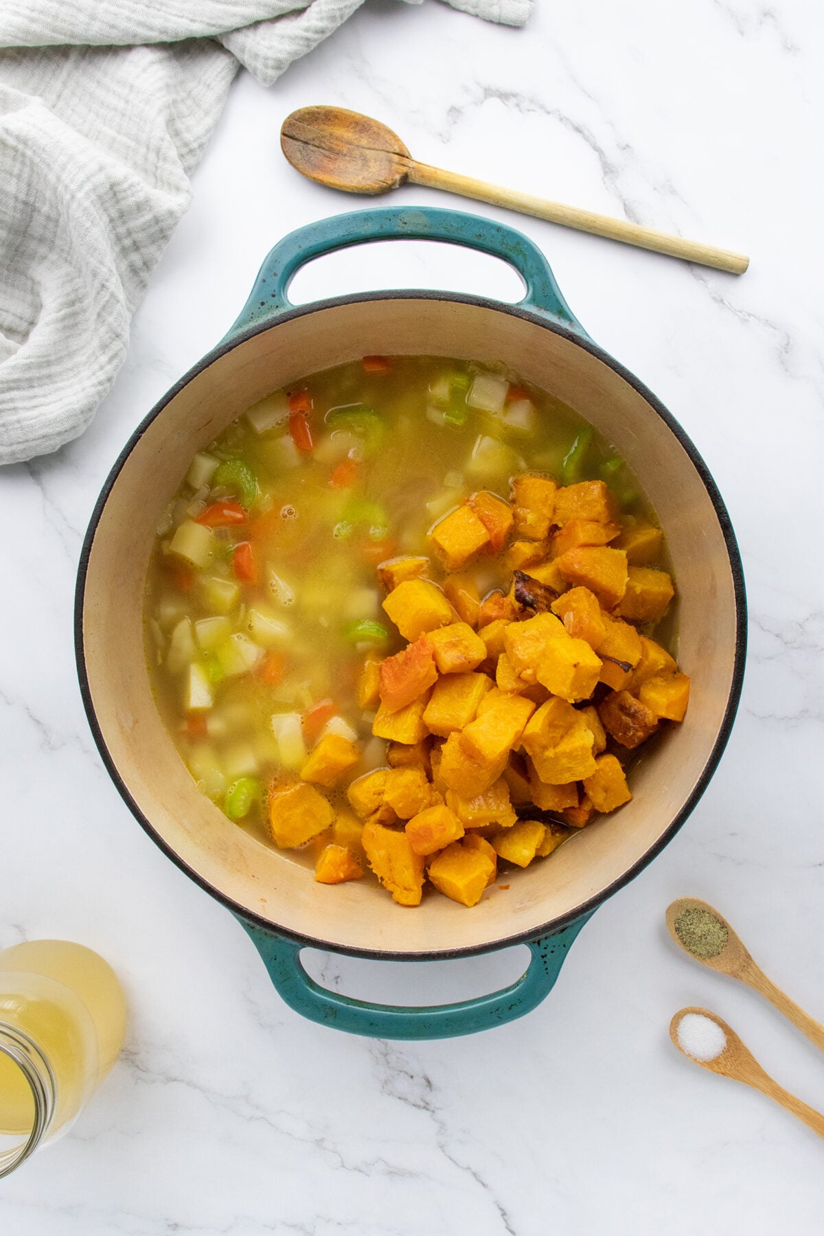A teal Dutch oven filled with soup topped with chunks of roasted butternut squash, surrounded by a wooden spoon, measuring spoons with spices, a jar of broth, and a striped cloth on a white marble surface.