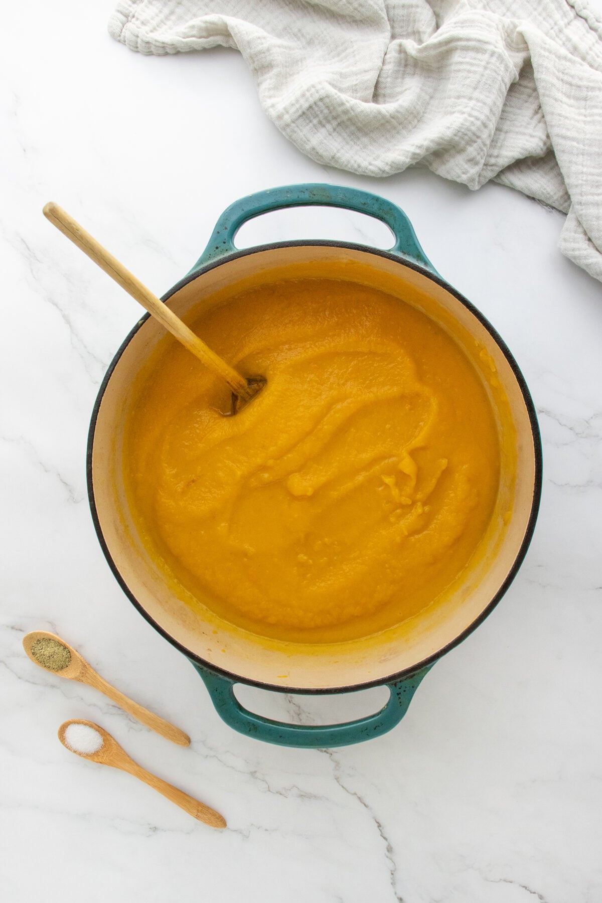A teal pot filled with creamy orange soup sits on a white marble surface, with a wooden spoon inside. Nearby are two wooden spoons, one with ground black pepper, and a light cloth is partially visible at the top.