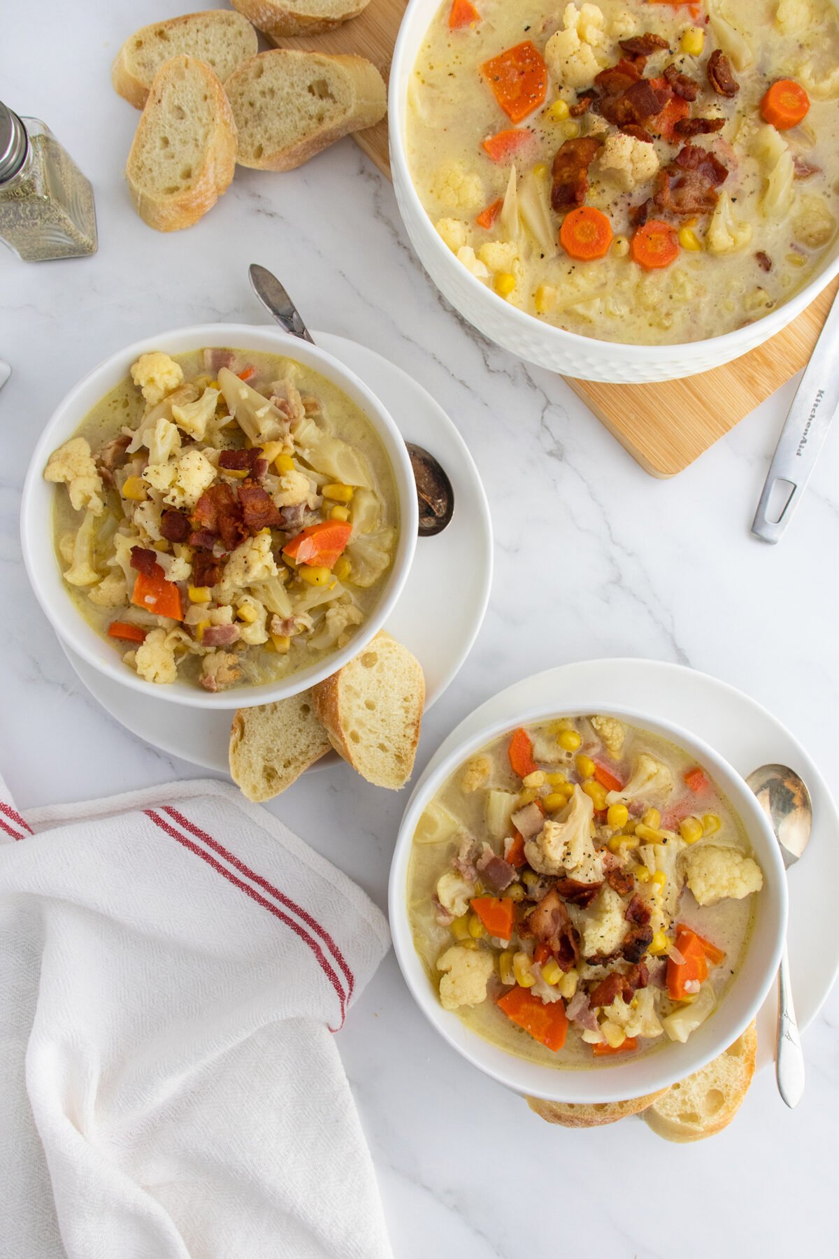 Bowls of hearty soup with vegetables, corn, and bacon are served with slices of crusty bread on a white marble table. Two spoons, a napkin, and a small breadboard are also visible.