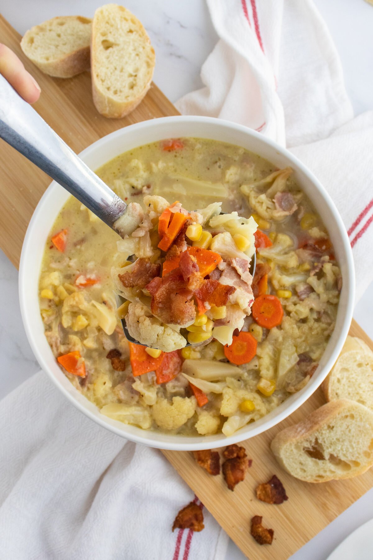 A bowl of creamy soup with carrots, cauliflower, corn, pasta, and bacon bits, served with slices of baguette on a wooden board. A spoon lifts a portion of the hearty soup above the bowl.