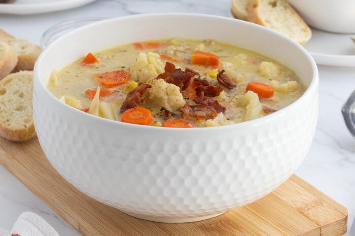 A white textured bowl filled with creamy soup containing cauliflower, carrots, pasta, and topped with crispy bacon sits on a wooden board with slices of bread nearby.