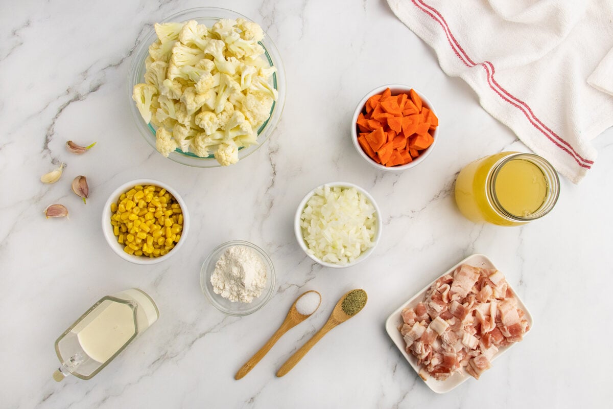 A top view of chopped cauliflower, carrots, onions, bacon, corn, garlic cloves, flour, black pepper, a jar of broth, a jug of cream, and a white towel on a marble countertop.