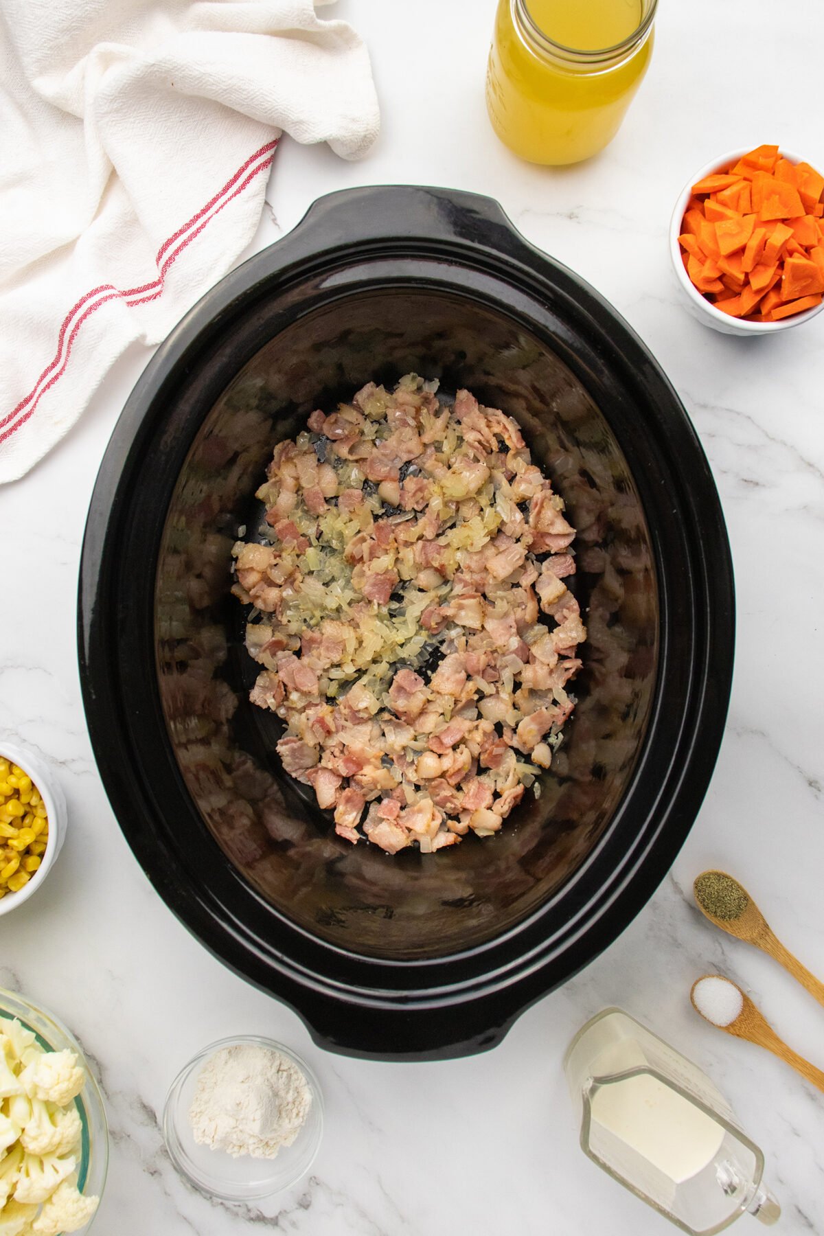 A slow cooker filled with chopped cooked chicken sits on a marble countertop, surrounded by bowls of chopped carrots, cauliflower, corn, flour, a jar of broth, a cup of milk, and measuring spoons with spices.