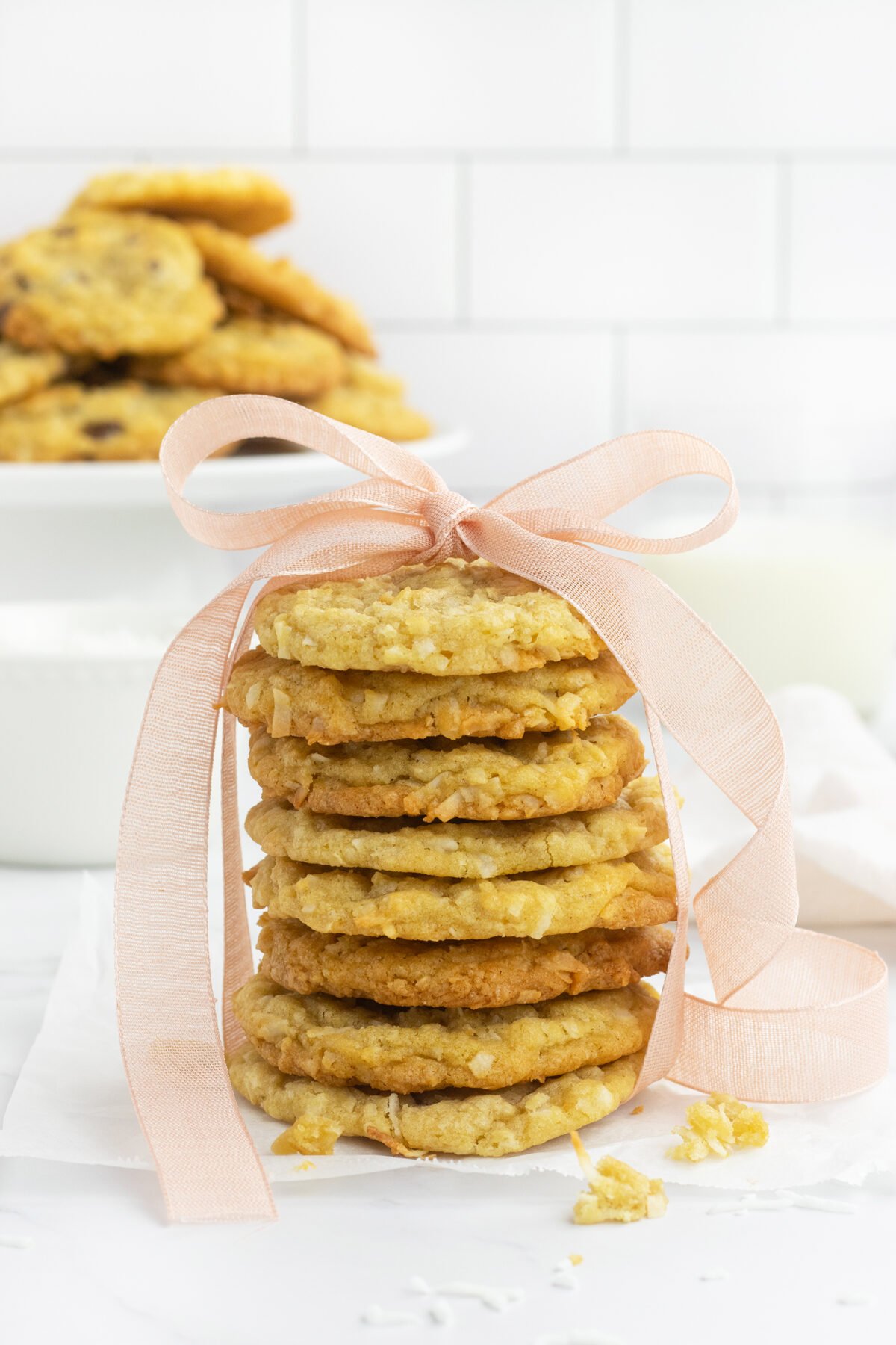 A stack of oatmeal cookies tied with a pale pink ribbon sits on parchment paper, with more cookies and a glass of milk in the blurred background.