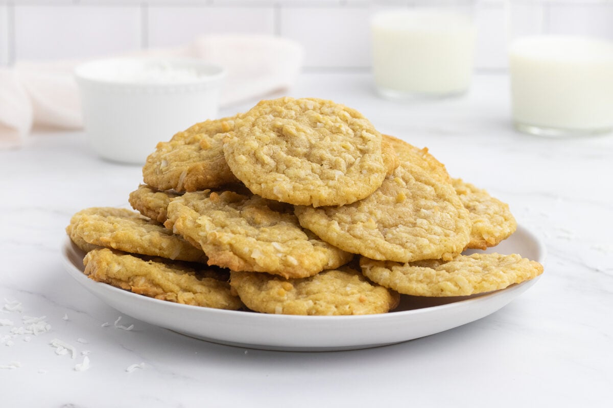 A white plate stacked with homemade oatmeal cookies on a marble surface, with blurred glasses of milk and a bowl in the background.