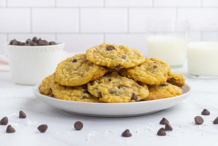 A plate of chocolate chip cookies sits on a white surface, with scattered chocolate chips and shredded coconut nearby. In the background, there are two glasses of milk and a bowl of chocolate chips.