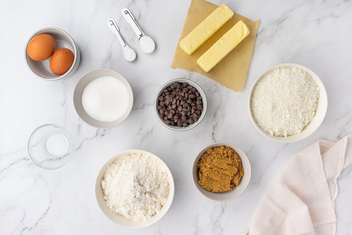 Baking ingredients on a marble surface, including eggs, butter sticks, chocolate chips, shredded coconut, flour, brown sugar, white sugar, salt, baking powder, and measuring spoons, with a cloth napkin in the corner.