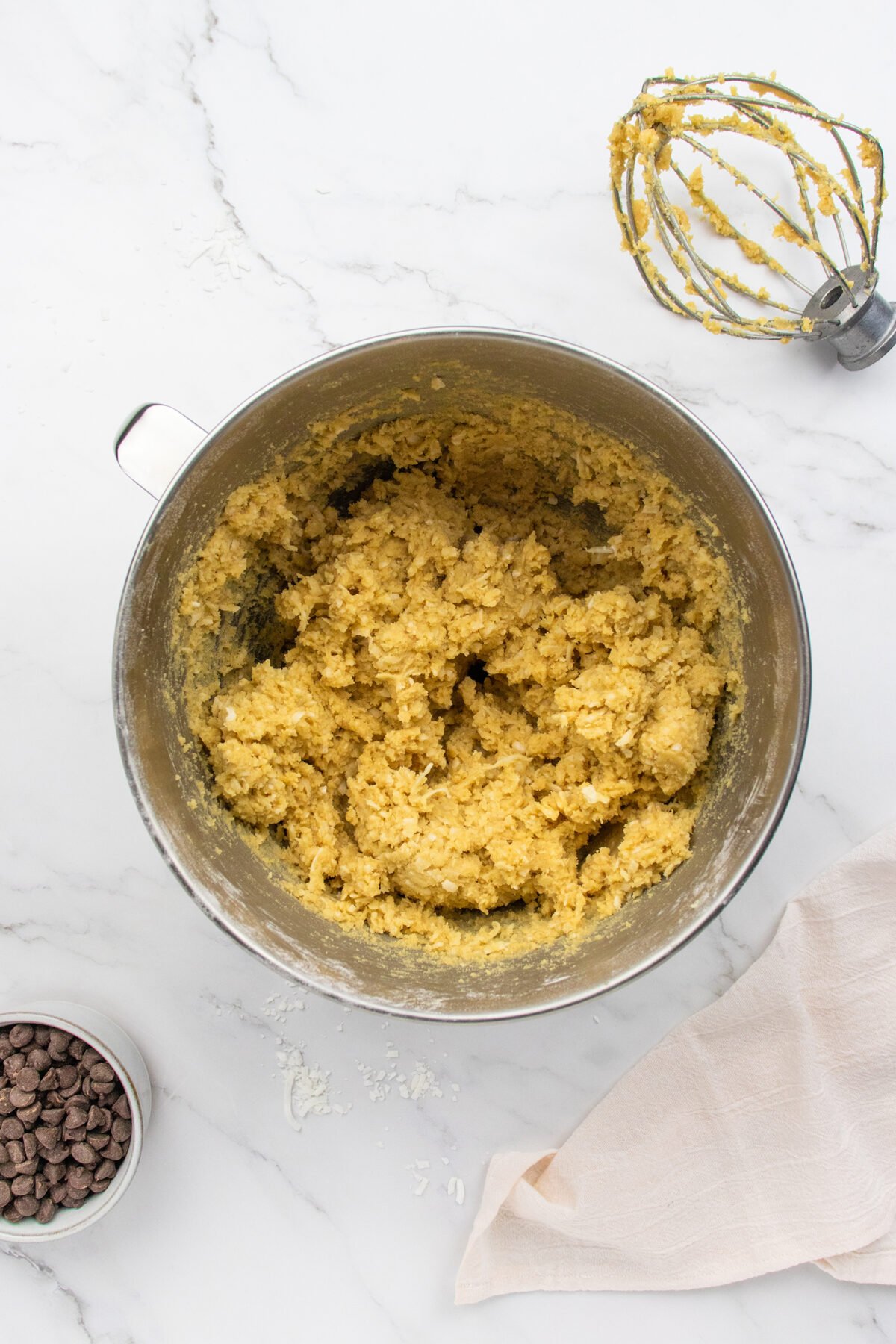 A mixing bowl filled with cookie dough sits on a white marble surface, surrounded by a whisk with dough on it, a piping bag, a small bowl of chocolate chips, and a light-colored cloth.