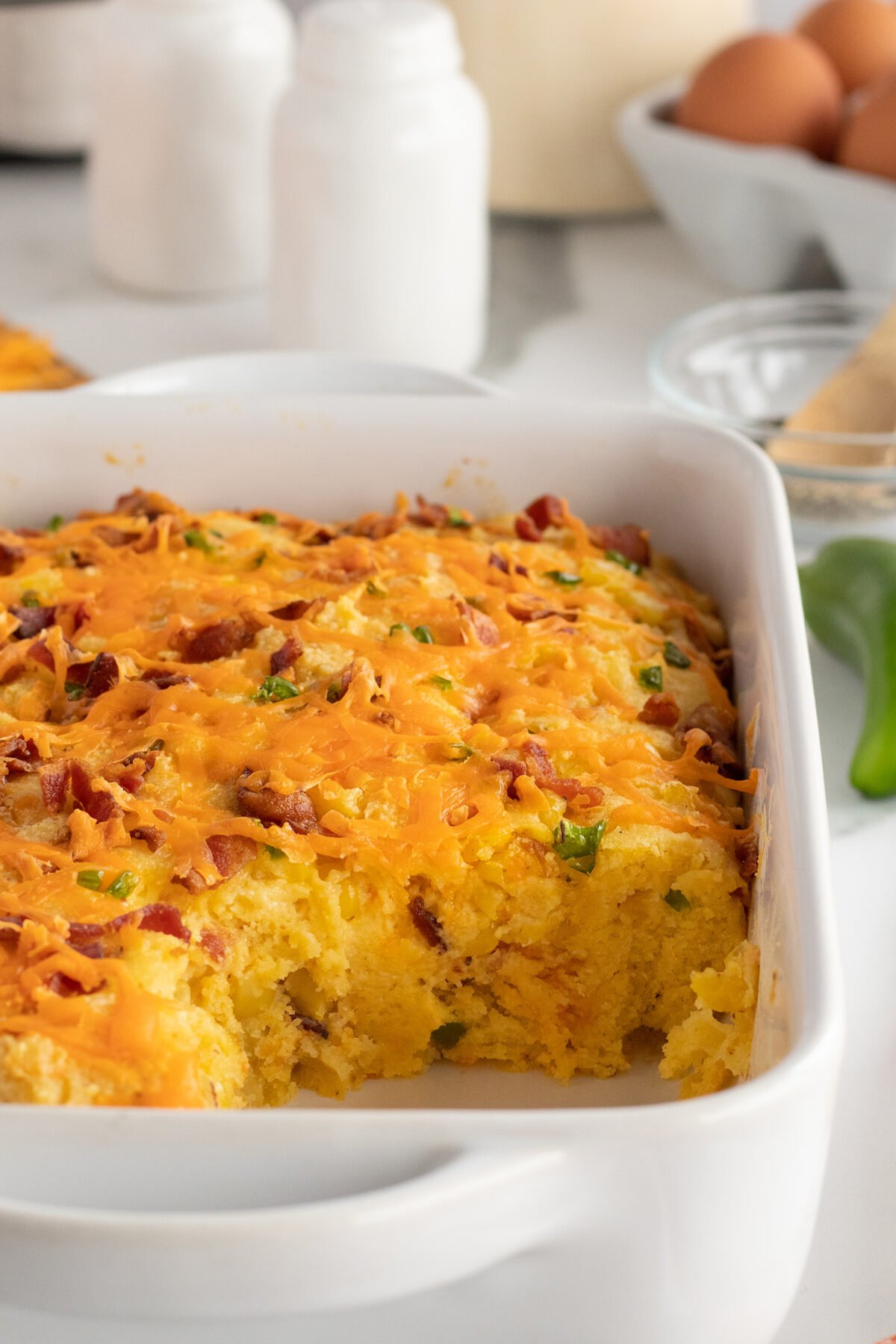A white casserole dish filled with cornbread topped with melted cheddar cheese, bits of bacon, and chopped green onions. A portion has been scooped out, showing the moist interior. Eggs and ingredients are in the background.