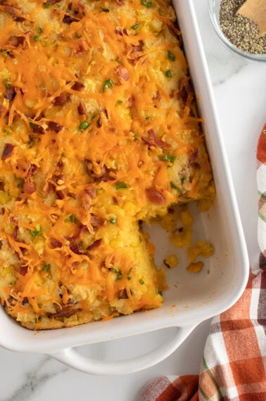A baked casserole topped with melted cheddar cheese, chopped green onions, and bacon bits, in a white dish with a corner piece missing. A bowl of pepper and a plaid towel are nearby on a marble surface.