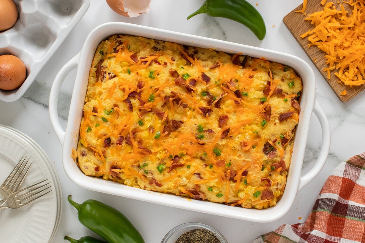 A baked breakfast casserole in a white dish, topped with melted cheese, bacon, and green bell peppers, surrounded by eggs, shredded cheese, forks, a plate, jalapeños, pepper, and a checkered napkin on a marble surface.