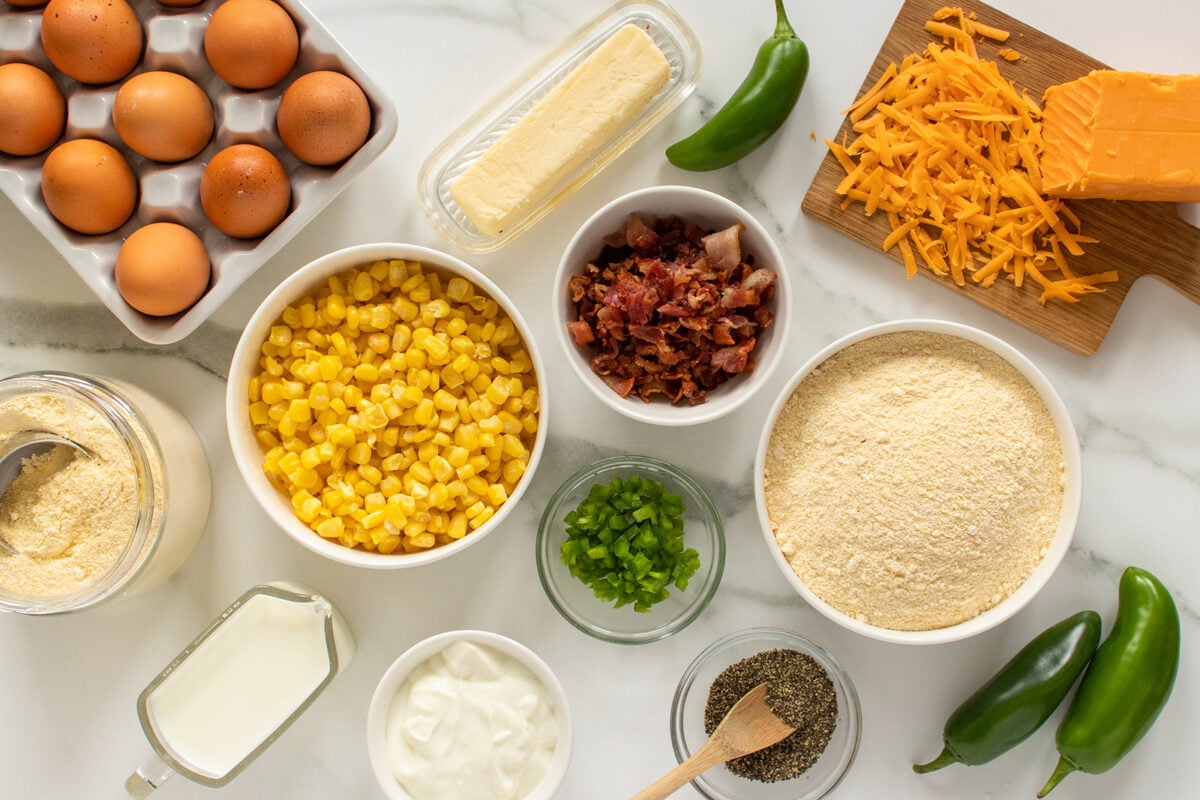 A top view of cornbread ingredients, including eggs, corn, flour, shredded cheese, chopped bacon, butter, sour cream, milk, chopped green onions, black pepper, and whole jalapeños arranged on a white surface.
