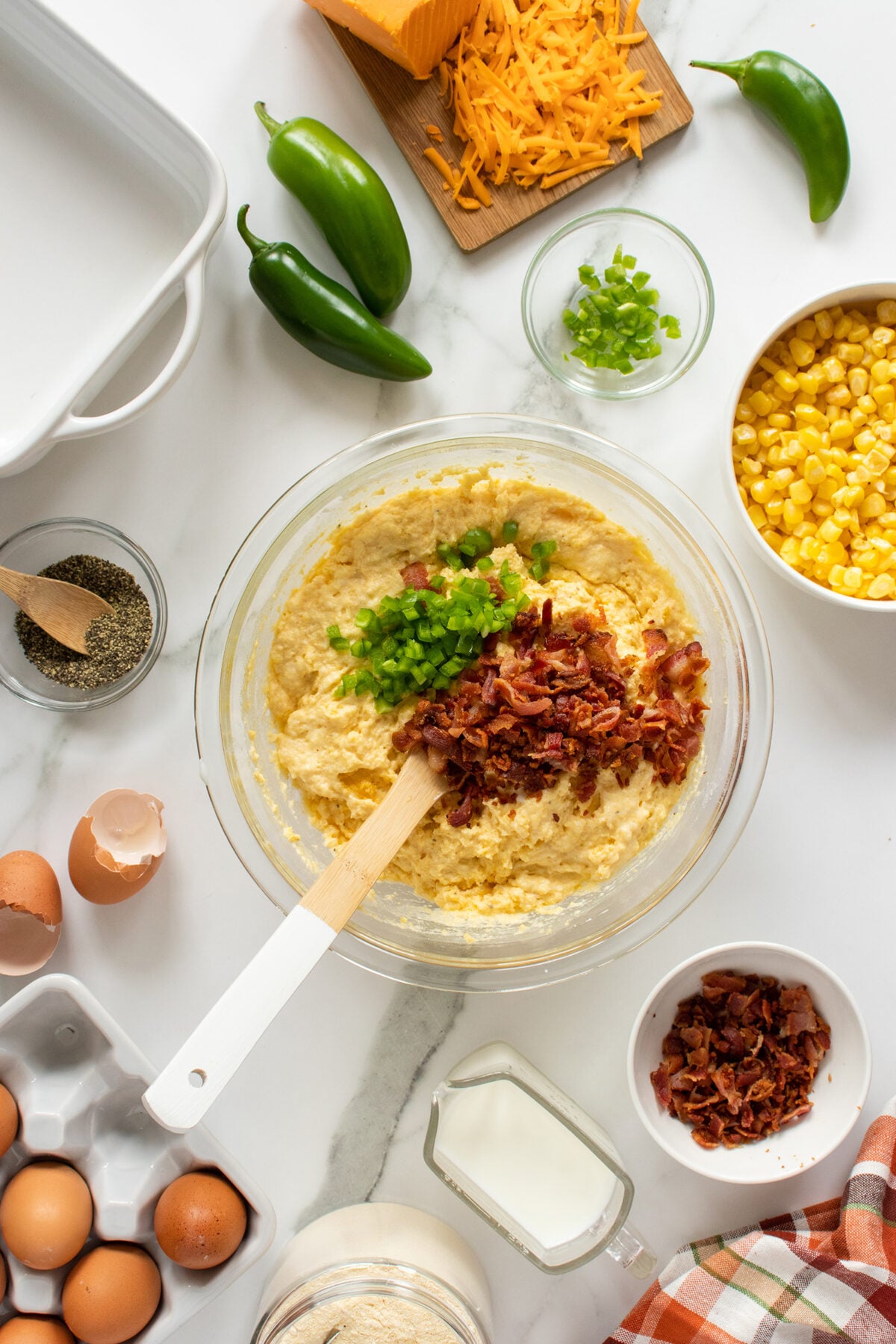 A glass bowl contains cornbread batter topped with chopped bacon and green peppers. Surrounding the bowl are eggs, milk, corn, shredded cheese, jalapeños, pepper, and a baking dish on a marble surface.