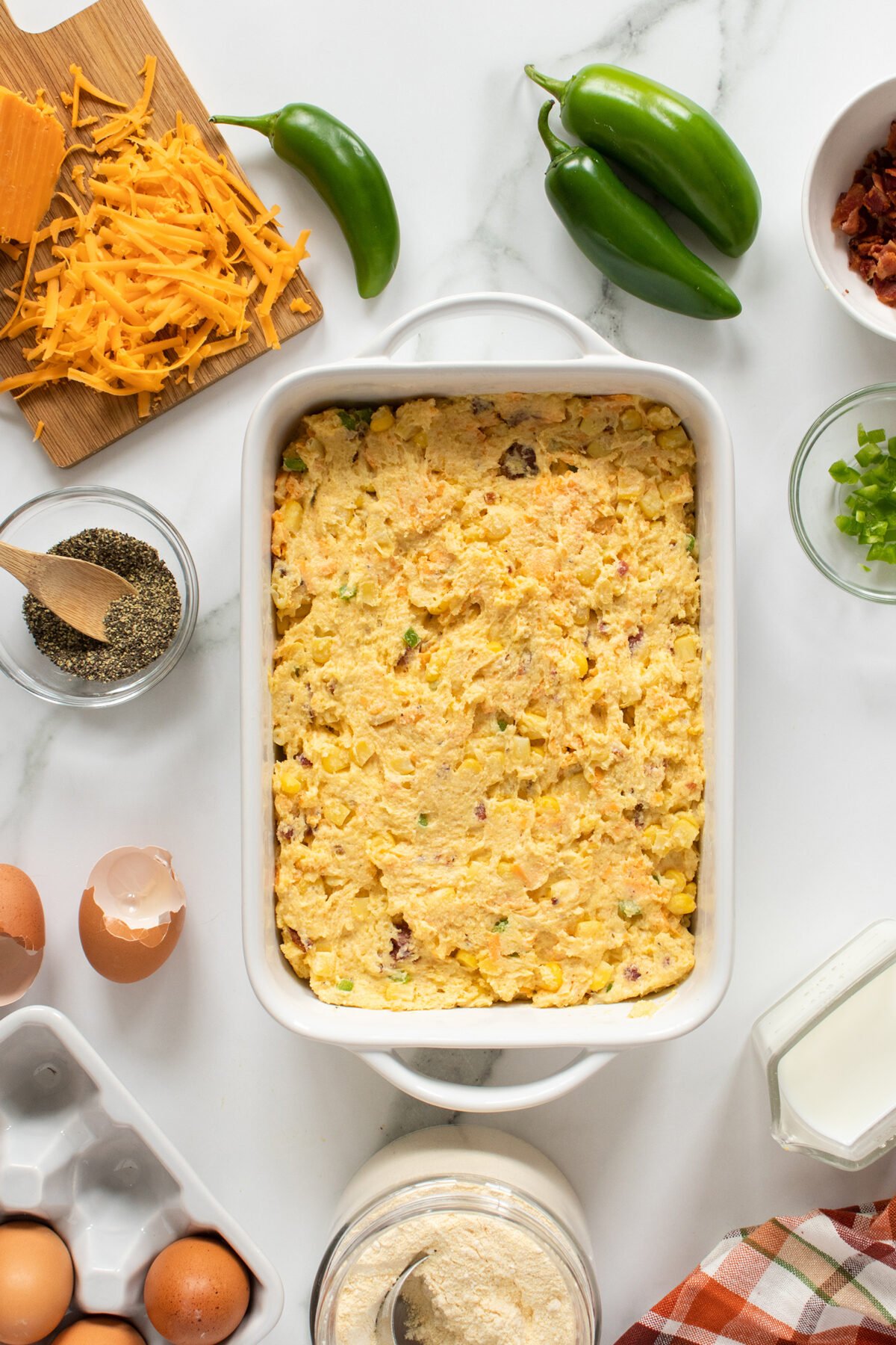 A casserole dish filled with a cheesy egg bake sits on a white countertop, surrounded by shredded cheese, eggs, jalapeños, green onions, bacon bits, ground pepper, milk, and a jar of flour.