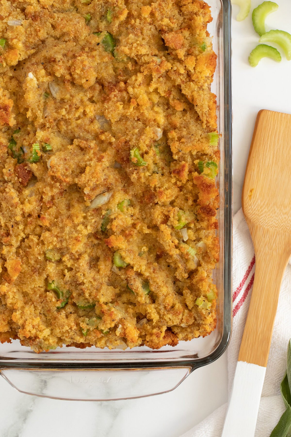 A glass baking dish filled with golden-brown cornbread stuffing, showing bits of celery. A wooden spatula and a white towel with a red stripe are nearby on a marble countertop.